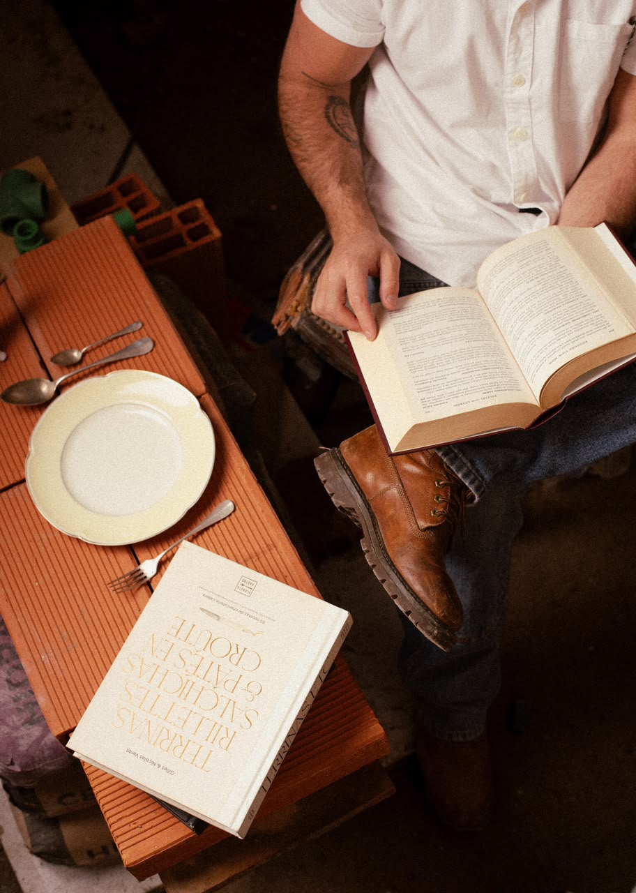 Vista desde arriba: un plato, cubiertos y libros de cocina sobre ladrillos; alguien lee sentado al lado.